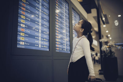 Cute girl looking arrival departure board while standing in airport