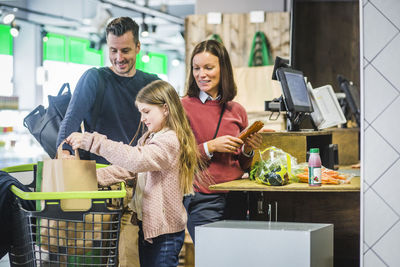 Smiling parents looking at daughter arranging paper bags in shopping cart