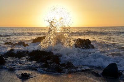 Waves splashing on rocks against sky during sunset