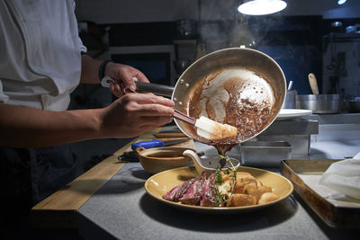 Side view of unrecognizable chef in uniform adding sauce in dish with slices of delicious beef placed on counter in kitchen