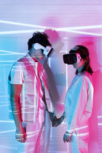 Side view of couple holding hands in modern vr goggles exploring cyberspace at wall with bright glowing lights in studio