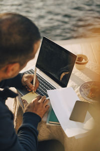 High angle view of mature man writing on adhesive note over laptop at table in patio