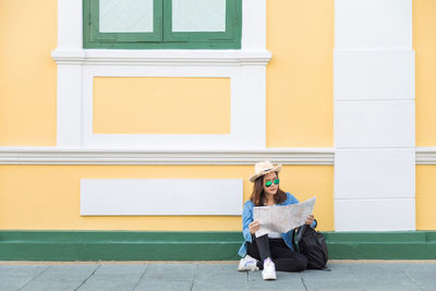 Full length of woman wearing sunglasses holding map while sitting against wall