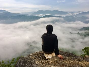 Rear view of man sitting at mountain edge in foggy weather