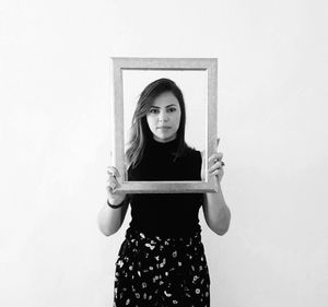 Portrait of young woman holding picture frame while standing against white background