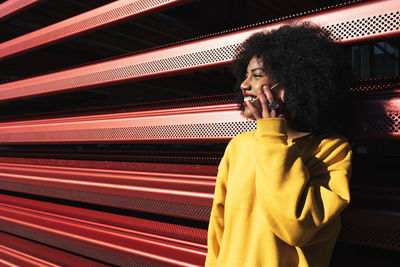 Smiling young woman listening over phone while standing metallic wall