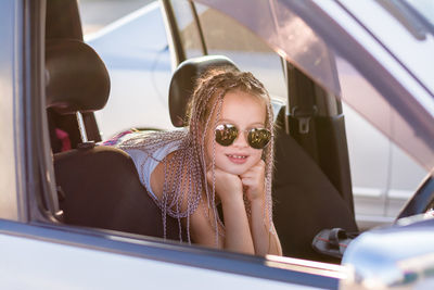 Smiling teenage girl with afro-braids sits in the passenger compartment of a car on a hot summer day