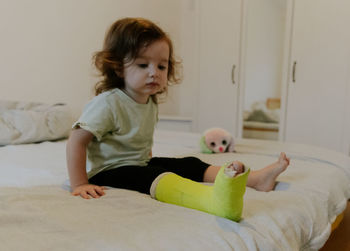 A happy girl with a cast on her leg is sitting on the bed.