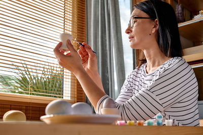 Woman preparing colored easter eggs and painting eggcup for easter holiday celebration.