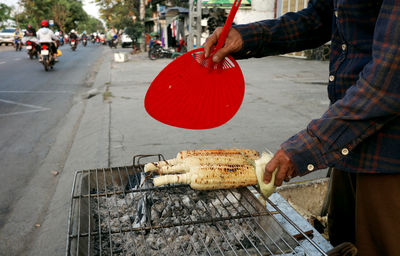 Close-up of the vendor's hand holding a fan over the grilled corn on the charcoal fire.