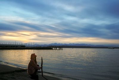 Side view of girl crouching with stick on shore at beach against sky during sunset