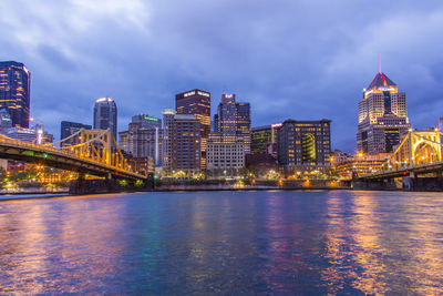 Bridges over river by illuminated buildings against sky at dusk