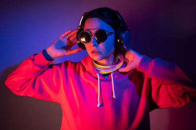 Cyberpunk woman in hooded hoodie and sunglasses dances against wall with neon sticks and headphone