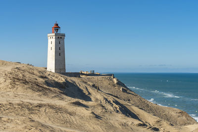 Lighthouse by sea against clear blue sky