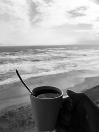 Close-up of hand holding coffee cup at beach against sky