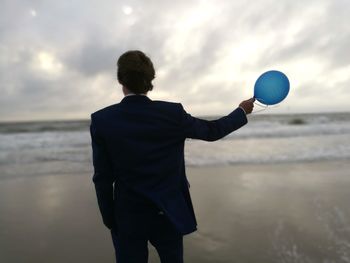Rear view of man wearing suit holding balloon while standing at beach against cloudy sky