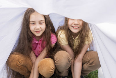 Two little girls, sisters with long hair sits under sheet on grass in meadow. caucasian asian