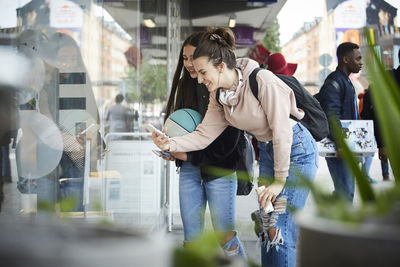 Smiling female friends photographing while standing by store window in city