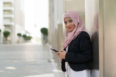 Portrait of smiling businesswoman wearing hijab using mobile phone while standing against building