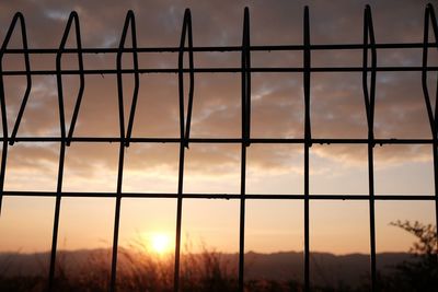 Silhouette metallic fence against cloudy sky during sunset