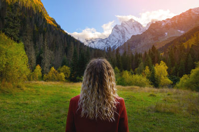 Rear view, a blonde woman in a red coat stands against the backdrop of snow-capped mountains and a