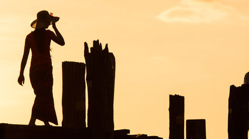 Silhouette woman standing by wooden post against sky during sunset