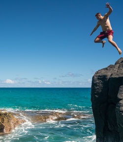 Low angle view of shirtless man jumping on rock at sea against sky