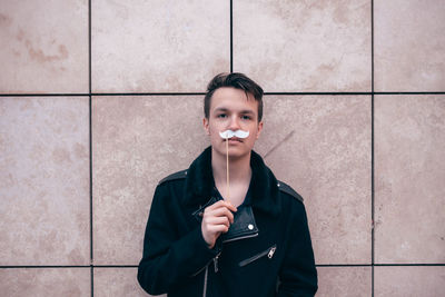 Portrait of young man holding mustache prop while standing against wall