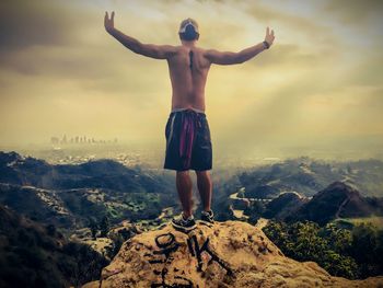 Rear view of shirtless man standing on mountain with arms outstretched against cloudy sky