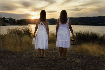 Rear view of friends holding lanterns while standing against lake during sunset