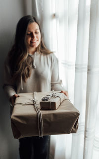 Smiling young woman holding gift box standing by window at home