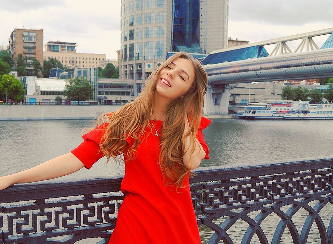 architecture, built structure, young adult, railing, water, one person, bridge - man made structure, real people, river, leisure activity, long hair, lifestyles, building exterior, outdoors, day, young women, smiling, beautiful woman, city, red, happiness, portrait, sky