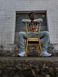 Full length of young man sitting on seat against brick wall