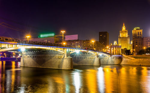 Illuminated bridge over river by buildings against sky at night