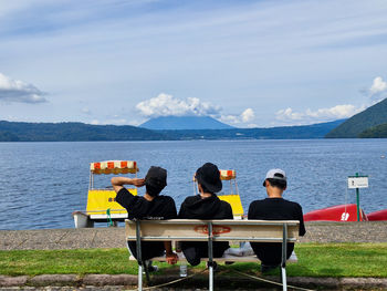 Rear view of people sitting on bench by sea against sky