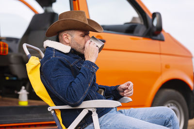 Man in hat drinking coffee while sitting on chair by motor home