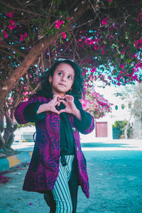 Girl making heart shape while standing against tree