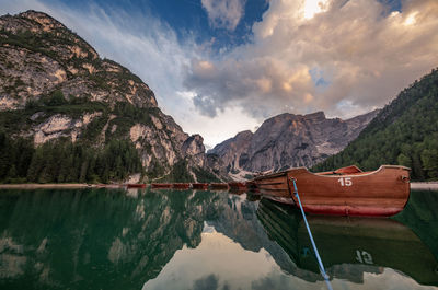 Boat moored on lake by mountains against cloudy sky during sunset