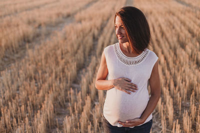 Smiling pregnant woman with hands on stomach standing on field