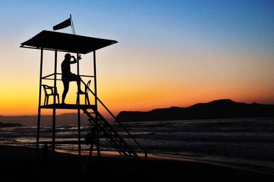 Full length of silhouette lifeguard on hut at beach against clear sky during sunset