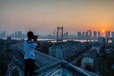 Rear view of man photographing bridge over river during sunset