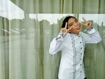 Portrait of playful girl showing peace sign while standing against window
