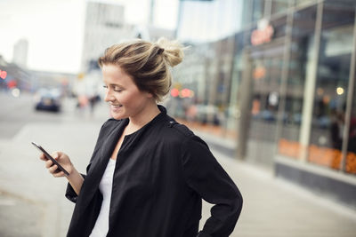 Happy woman holding mobile phone while standing in city