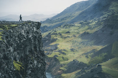 Man walking on cliff by mountains against sky