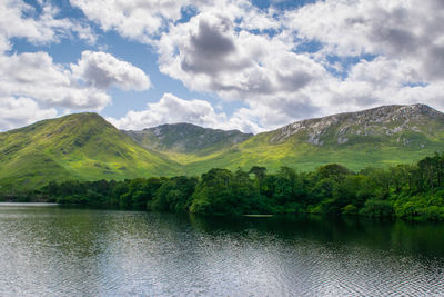 Scenic view of green mountains by lake against cloudy sky