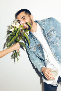 Portrait of smiling young man smelling flowers held by woman against white wall at home