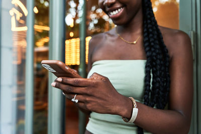 Crop african american female in stylish top smiling and sending text message on smartphone while leaning on glass wall on city street