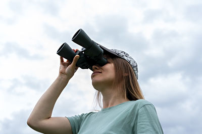 Young blonde woman bird watcher looking through binoculars at cloudy sky ornithological research 