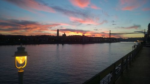 Illuminated city by river against sky during sunset