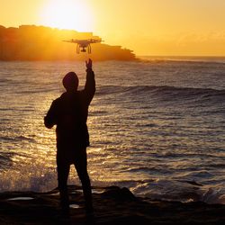 Rear view of silhouette man controlling quadcopter at beach against sky